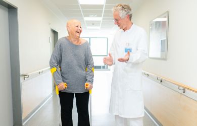 Eine Patientin steht mit Unterarmgehstützen auf einem Klinikflur und lacht im Gespräch mit einem Arzt im weißen Kittel.