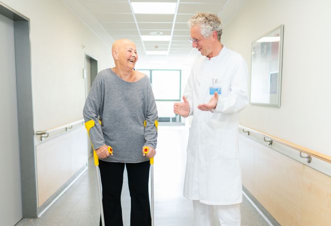 Eine Patientin steht mit Unterarmgehstützen auf einem Klinikflur und lacht im Gespräch mit einem Arzt im weißen Kittel.