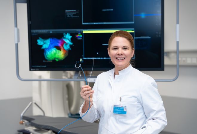 Eine Ärztin im weißen Kittel steht in einem Behandlungsraum und hält einen Katheter in der Hand. Sie blickt lächelnd in die Kamera. Hinter ihr ist ein Monitor mit einer farbigen medizinischen Darstellung zu sehen.