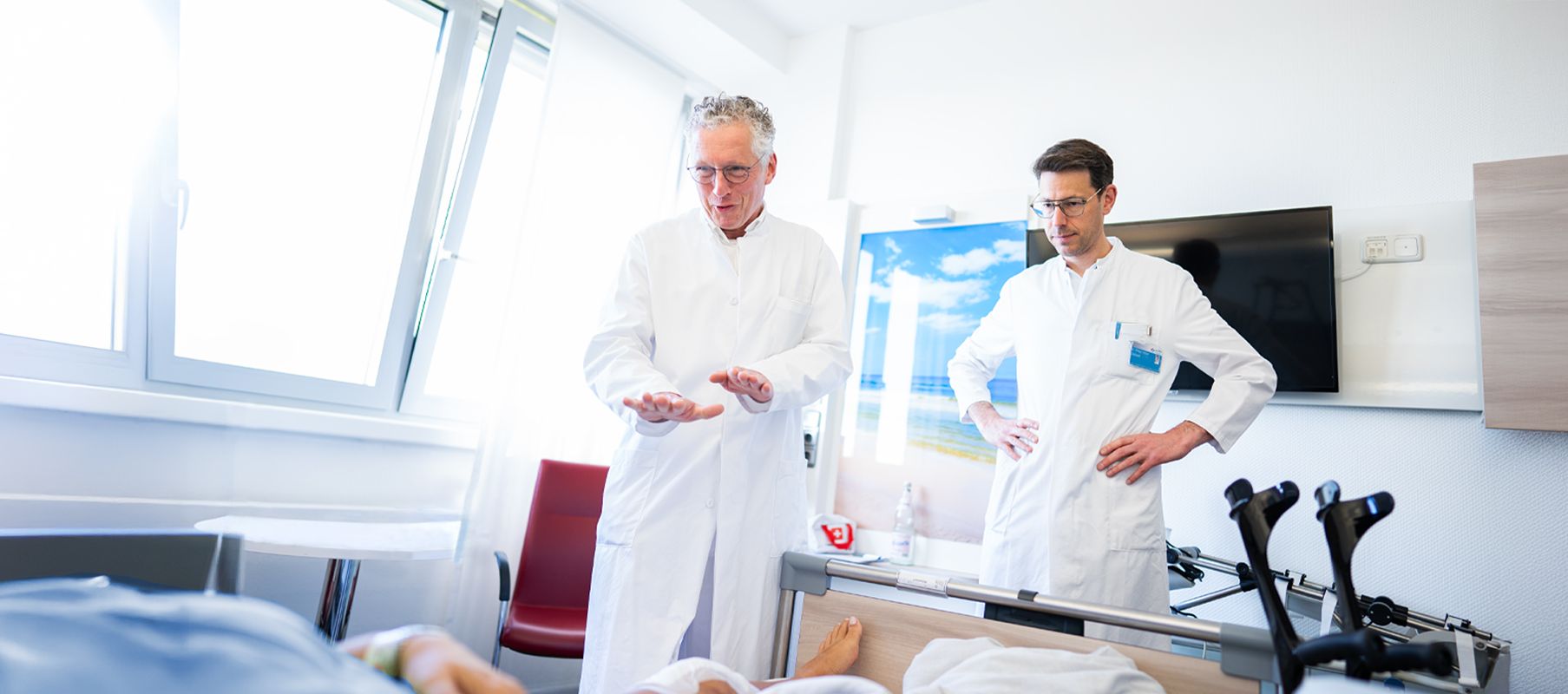 Zwei Ärzte in weißen Kitteln stehen in einem Patientenzimmer neben einem Krankenhausbett und sprechen mit einer liegenden Person. Am Bett stehen rechts zwei Gehkrücken.