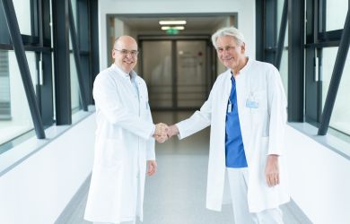 Prof. Dr. Schöls (rechts) und Prof. Jánosi (links) reichen sich im Klinikflur mit großen Fenstern die Hand. Beide tragen Kittel und wirken sehr kollegial.