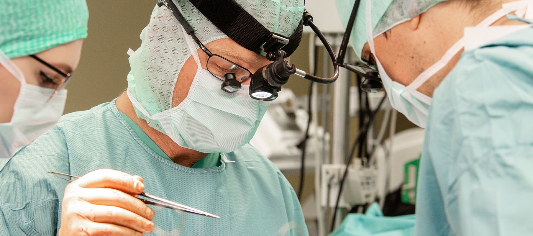 In einem modernen Operationssaal steht Professor Börgermann gemeinsam mit einer OP-Schwester und einem weiteren Chirurgen an einem Operationstisch. Alle drei tragen sterile grüne OP-Kleidung, einschließlich Mundschutz, Haube und Handschuhen.