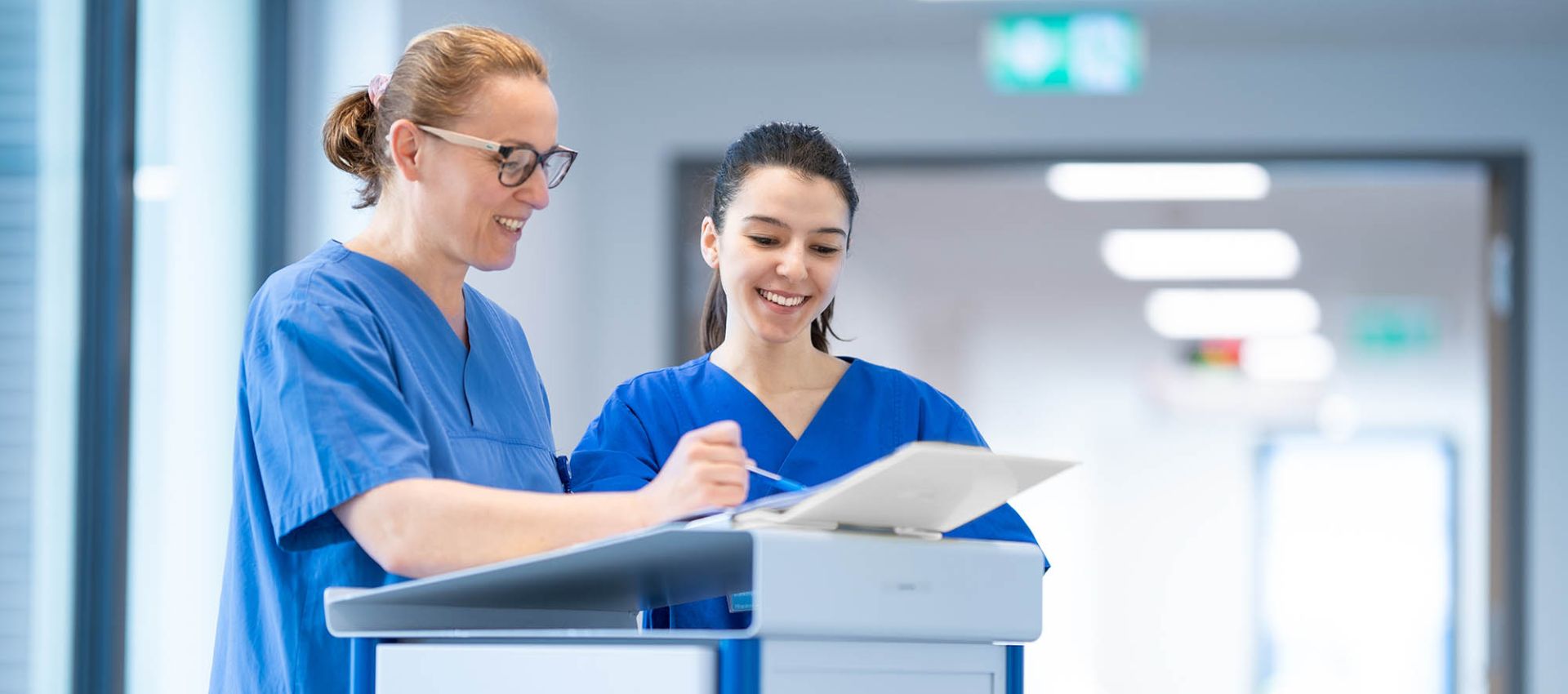 Zwei weibliche Pflegekräfte stehen im Stationsflur an einem Dokumentationswagen und schauen sich gemeinsam Patientendaten an.