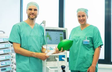 Zwei Anästhesist*innen in grüner OP-Kleidung stehen in einem Behandlungsraum vor einem Beatmungsgerät. Dr. Steffen Veen hält einen grünen Beatmungsball, Dr. Stefanie Klenke steht daneben und lächelt.