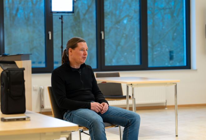 Ein Mann sitzt auf einen Stuhl in einem großen, hellen freundlichen Raum. Im Hintergrund ist eine große Fensterfront zu sehen. Hinter ihm steht ein Scheinwerfer. Er ist im Gespräch.