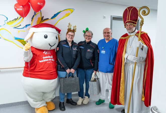 Gruppenfoto mit dem Maskottchen Eva im Weihnachtsoutfit,  zwei Malteser-Mitarbeiterinnen mit festlichem Haarschmuck, Dr. Tarusinov aus der Kinderkardiologie und dem Nikolaus der Malteser, aufgenommen im Krankenhausflur vor einer bunt bemalten Wand.