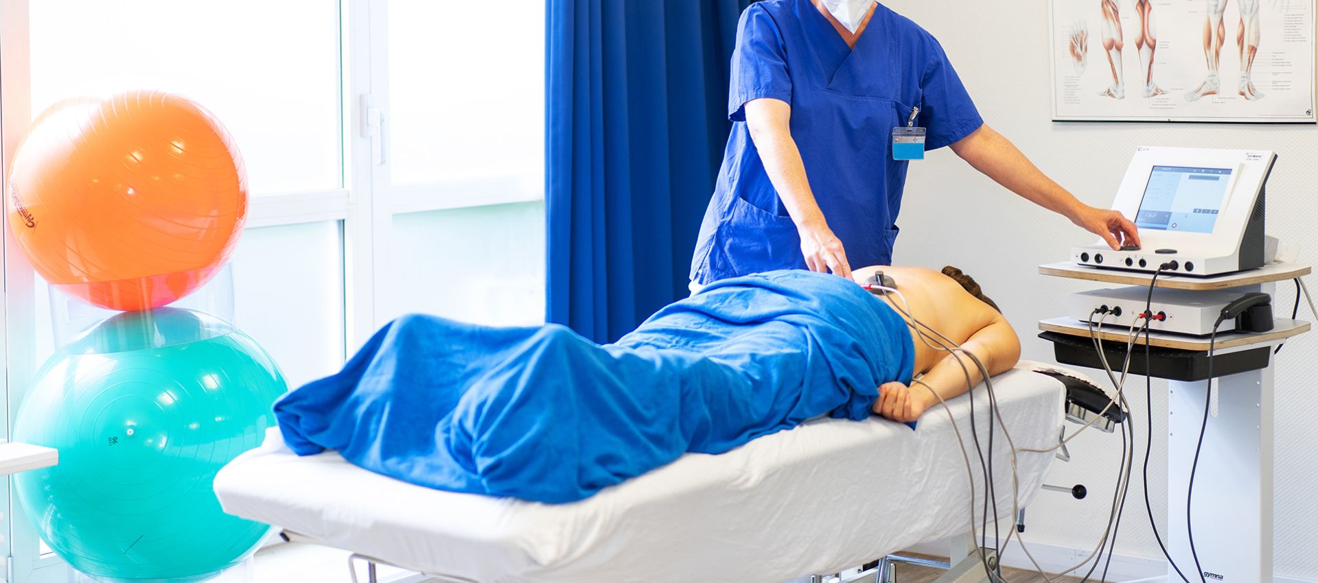 Physiotherapeutin bedient ein Gerät, während eine Patientin mit Therapie-Pads auf dem Rücken auf dem Bauch auf einer Liege liegt.