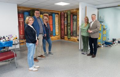 Mehrere Personen stehen im Klinikflur vor einer fertig gestalteten Wand mit großformatigen, bunten Bücher-Motiven. Zwei der beteiligten Mitarbeiterinnen stehen daneben, Malutensilien sind im Hintergrund zu sehen.