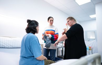 In einem Übungsraum der Pflegeschule demonstriert eine Ausbilderin die Blutdruckmessung. Eine Auszubildende sitzt auf dem Pflegebett, eine weitere beobachtet aufmerksam. Die Szene zeigt eine praktische Lernsituation im Unterricht.