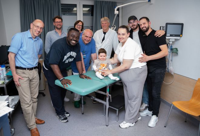 Mehrere Erwachsene und ein Kleinkind stehen gemeinsam in einem Behandlungsraum des Kinderherzzentrums und lächeln in die Kamera. Das Kind sitzt auf einer Untersuchungsliege und hält ein Kuscheltier im Arm.