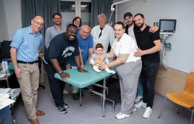 Mehrere Erwachsene und ein Kleinkind stehen gemeinsam in einem Behandlungsraum des Kinderherzzentrums und lächeln in die Kamera. Das Kind sitzt auf einer Untersuchungsliege und hält ein Kuscheltier im Arm.