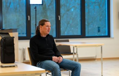 Ein Mann sitzt auf einen Stuhl in einem großen, hellen freundlichen Raum. Im Hintergrund ist eine große Fensterfront zu sehen. Hinter ihm steht ein Scheinwerfer. Er ist im Gespräch.