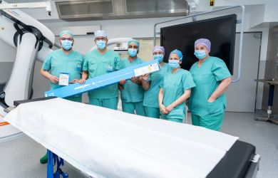 Mehrere Mitarbeitende eines OP-Teams stehen in grüner Schutzkleidung und mit Mundschutz in einem Eingriffsraum neben einem Behandlungstisch. Zwei Personen halten die Verpackung eines medizinisches Produktes in den Händen.