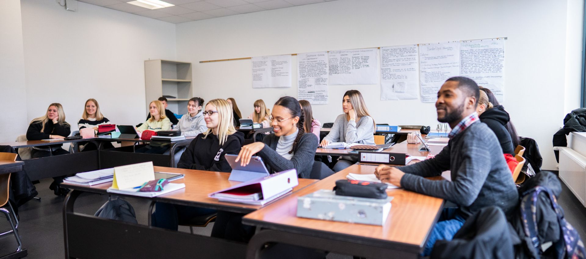 Auszubildende sitzen in einem freundlichen, lichtdurchfluteten Klassenraum der Pflegeschule. Sie arbeiten an ihren Plätzen, eine Lehrkraft ist nicht zu sehen. Die Szene vermittelt konzentriertes, eigenständiges Lernen.