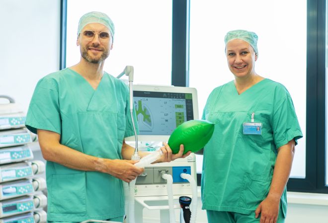 Zwei Anästhesist*innen in grüner OP-Kleidung stehen in einem Behandlungsraum vor einem Beatmungsgerät. Dr. Steffen Veen hält einen grünen Beatmungsball, Dr. Stefanie Klenke steht daneben und lächelt.