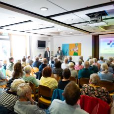 Zahlreiche Besucherinnen und Besucher sitzen in einem Veranstaltungsraum. Sie sitzen in Stuhlreihen nebeneinander und sind von hinten zu sehen. Sie verfolgen einen Vortrag mit Präsentation.