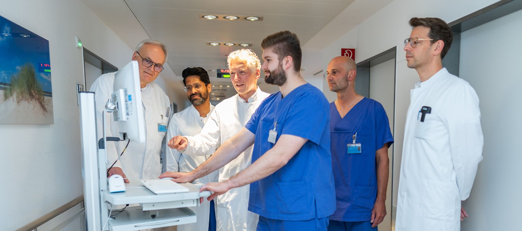 Mehrere Ärzte stehen gemeinsam auf einem Klinikflur an einem mobilen Computerwagen und besprechen einen Patientenfall. Einige tragen weiße Arztkittel, andere blaue OP-Kleidung.