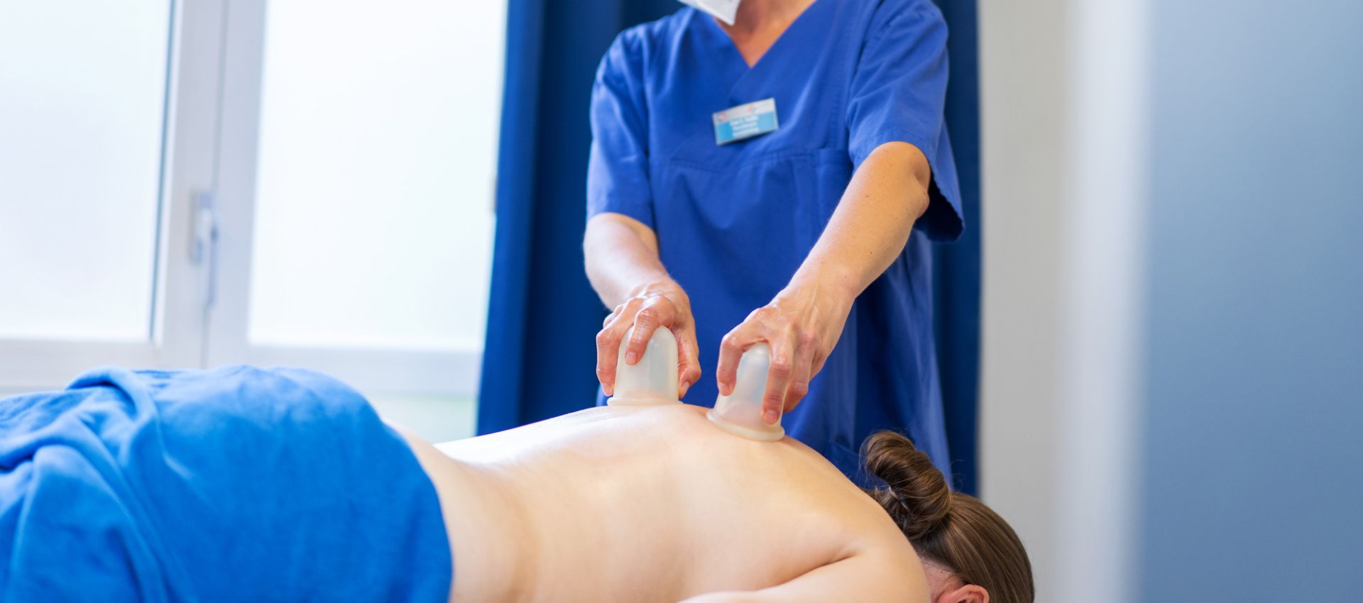 Eine Physiotherapeutin führt eine Schröpfmassage mit zwei Saugnäpfen am Rücken einer Patientin durch, die auf dem Bauch auf einer Therapieliege liegt.