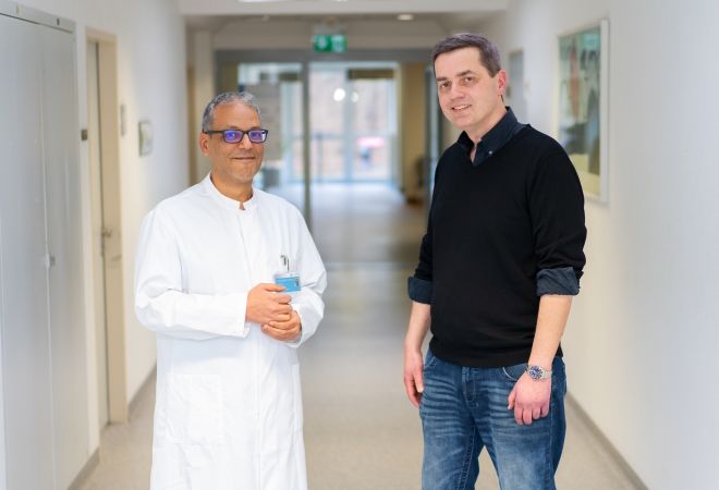 Der Patient Dino Andree Rogge steht gemeinsam mit Dr. Rashad El-Habony, leitender Oberarzt der Neurochirurgie, in einem hellen Klinikflur des Evangelischen Klinikums Niederrhein. Beide blicken in die Kamera und lächeln.