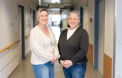 Die Pain Nurses Corinna Noy und Annika Trapphoff stehen nebeneinander in einem Krankenhausflur und lächeln in die Kamera.