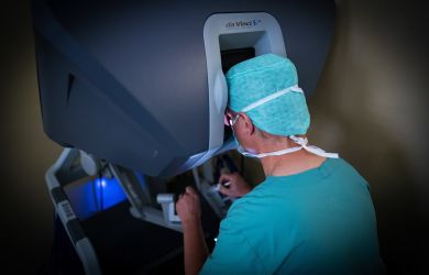 Prof. Dr. Börgermann in OP-Kleidung sitzt an der Steuerkonsole des Da-Vinci-Operationssystems und blickt durch das Okular. Aufnahme von hinten während eines Eingriffs.
