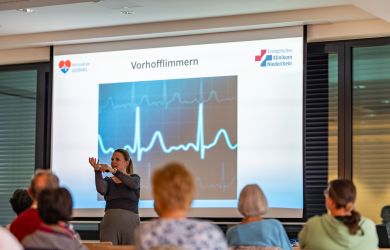 Dr. med. Ute Ruprecht steht vor einem Publikum und hält einen Vortrag über Vorhofflimmern. Hinter ihr ist eine große Präsentation mit einer Herzkurve zu sehen.