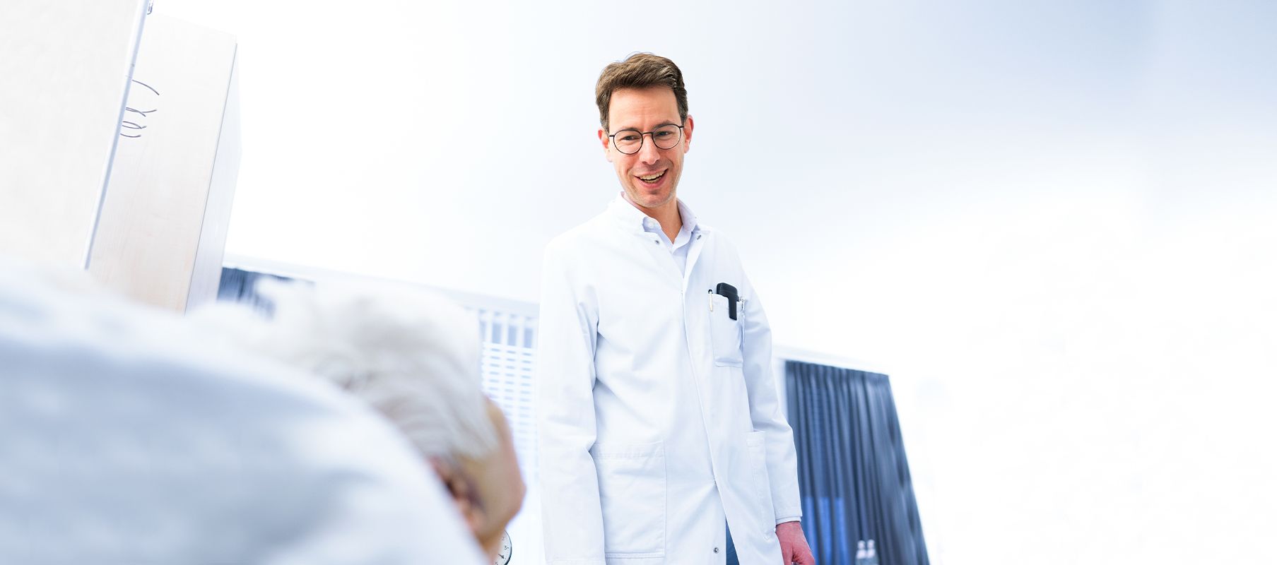 Dr. Philipp Weber steht in einem Krankenhauszimmer neben einem Patientenbett und spricht mit einer Patientin. Er trägt einen weißen Arztkittel.