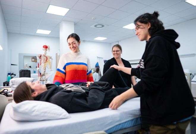 In einem Übungsraum der Pflegeschule liegt eine Auszubildende auf einem Pflegebett, während ihre Mitlernenden eine Pflegetechnik üben. Eine Ausbilderin begleitet die Situation lächelnd. Im Hintergrund ist ein Skelettmodell sichtbar.