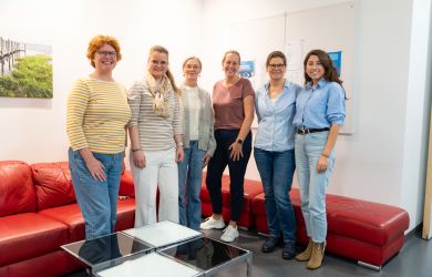 Sechs Frauen stehen nebeneinander in einem Raum vor einer roten Couch und lächeln in die Kamera. Es handelt sich um Lehrerinnen aus Finnland, Vertreterinnen des Walther-Rathenau-Berufskollegs sowie die stellvertretende Leitung der Pflegeschule am EVKLN.