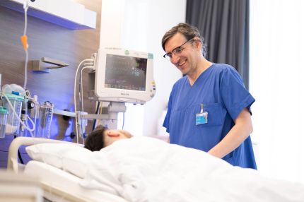 Eine männliche Pflegekraft steht am Patientenbett, lächelt dem Patienten zu und begleitet eine Pflegesituation. Im Hintergrund ist ein Monitor mit Vitalzeichen sichtbar.