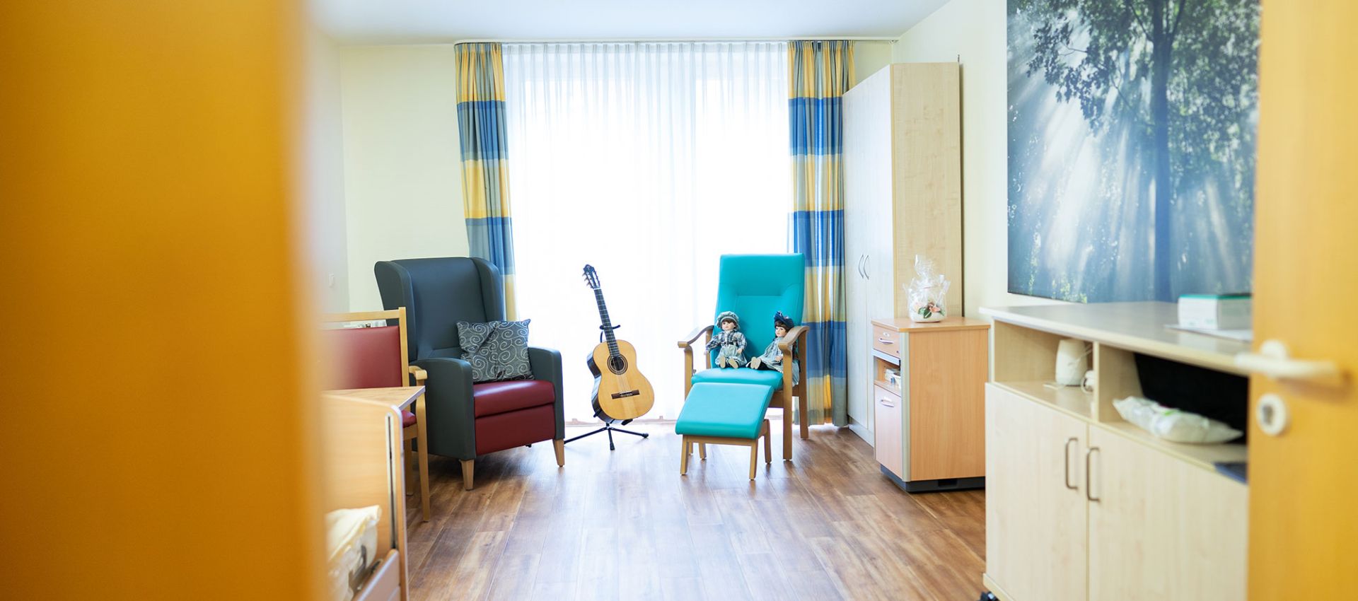 Blick durch die geöffnete Tür in die Ruheoase: Zwei gemütliche Sessel, dazwischen steht eine Gitarre. Ein großes Fenster mit Vorhängen lässt Tageslicht in den Raum.