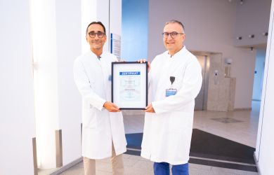 Zwei Ärzte in weißen Kitteln stehen nebeneinander in einem hell beleuchteten Krankenhausflur. Gemeinsam halten sie ein gerahmtes Zertifikat in die Kamera und lächeln.