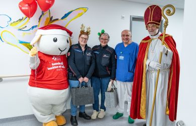 Gruppenfoto mit dem Maskottchen Eva im Weihnachtsoutfit,  zwei Malteser-Mitarbeiterinnen mit festlichem Haarschmuck, Dr. Tarusinov aus der Kinderkardiologie und dem Nikolaus der Malteser, aufgenommen im Krankenhausflur vor einer bunt bemalten Wand.