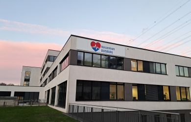 Außenansicht des Herzzentrums Duisburg am Standort in Duisburg Fahrn. An der Fassade ist das Logo des Herzzentrums angebracht. Der Himmel im Hintergrund ist rosa vom Sonnenaufgang.