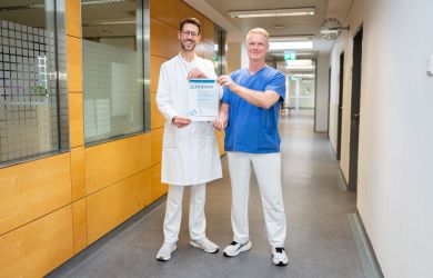 Dr. Philipp Weber (links) und Kersten Brenner (rechts) stehen lächelnd in einem Krankenhausflur und halten gemeinsam das Zertifikat des Regionalen Traumazentrums in die Kamera. Weber trägt einen weißen Arztkittel, Brenner blaue OP-Kleidung.