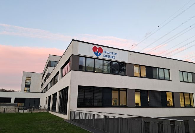 Außenansicht des Herzzentrums Duisburg am Standort in Duisburg Fahrn. An der Fassade ist das Logo des Herzzentrums angebracht. Der Himmel im Hintergrund ist rosa vom Sonnenaufgang.