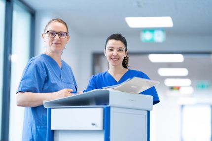 Zwei weibliche Pflegekräfte dokumentieren im Stationsflur gemeinsam Patientendaten an einem Dokumentationswagen.