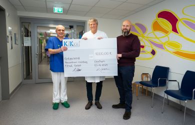 Dr. Gleb Tarusinov aus der Kinderkardiologie, Dr. Michael Scheid (Kinderherzchirurgie) und Marcel Radomski (KiKxxL) stehen im Krankenhausflur und halten gemeinsam einen großen symbolischen Spendencheck über 1000 Euro.