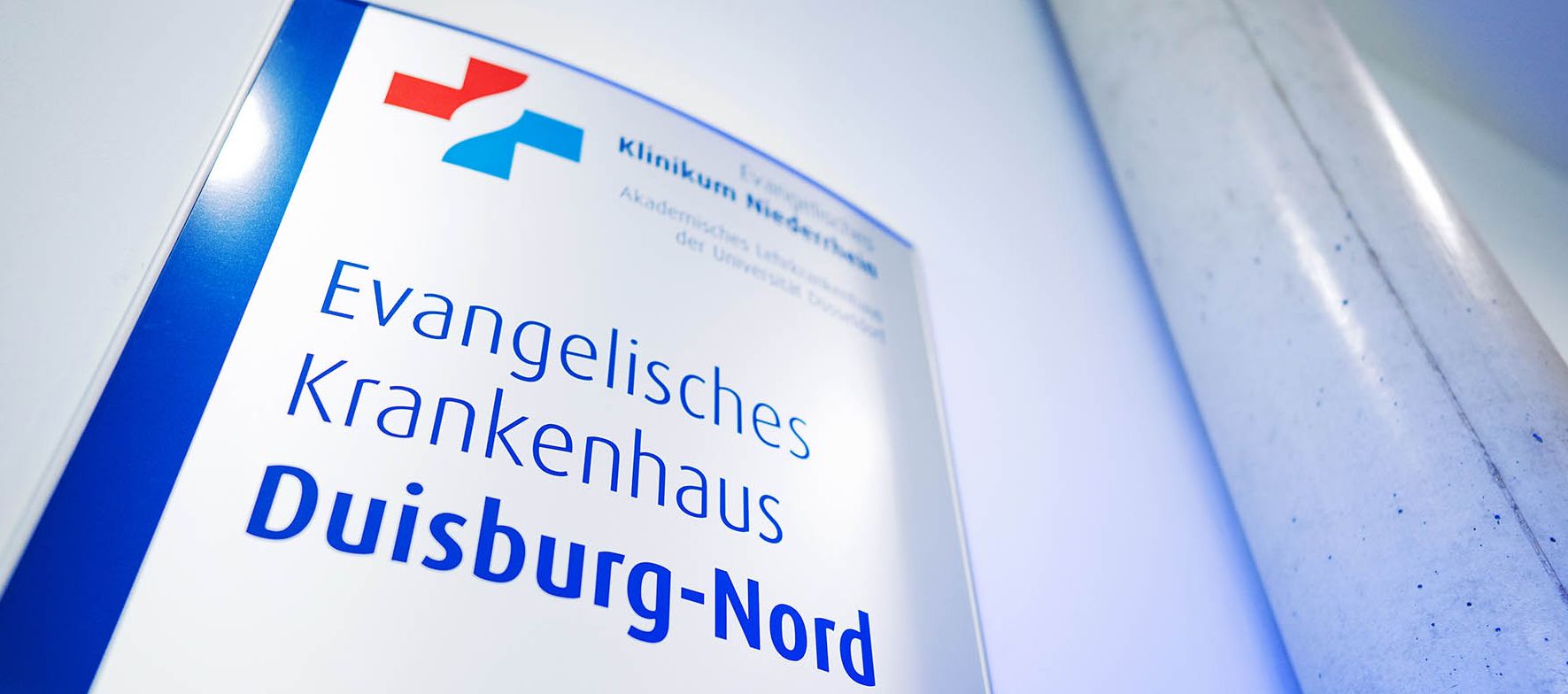 Ein Schild mit dem Namen des Evangelischen Krankenhauses Duisburg-Nord hängt gut sichtbar im Eingangsbereich. Es zeigt auch das rot-blaue Kreuz als Logo des Klinikverbundes.