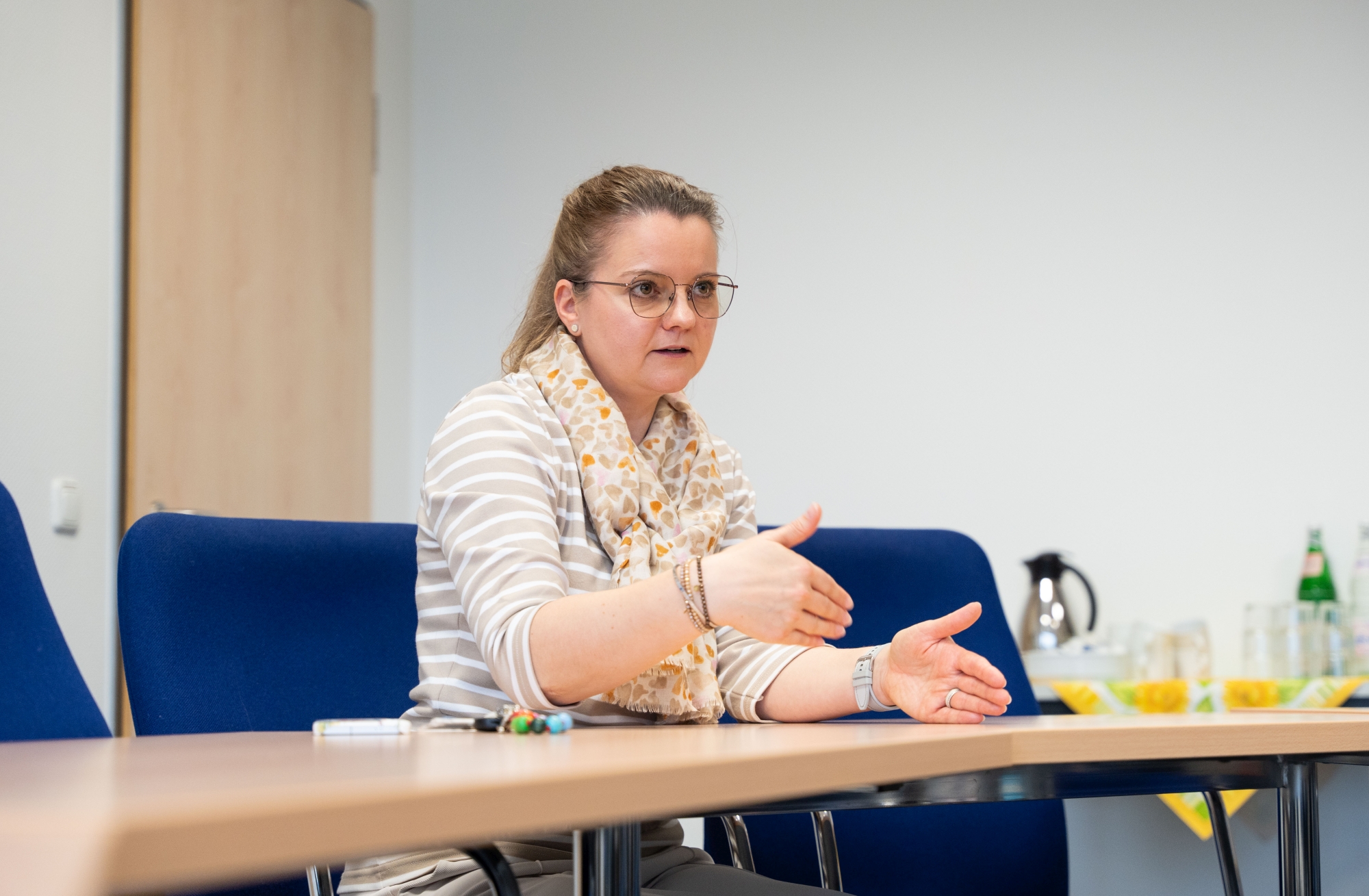 Sonja Armbruster, stellvertretende Leitung der Pflegeschule am EVKLN, sitzt an einem Tisch und spricht gestikulierend. Vor ihr liegen Stifte, im Hintergrund stehen Getränke.