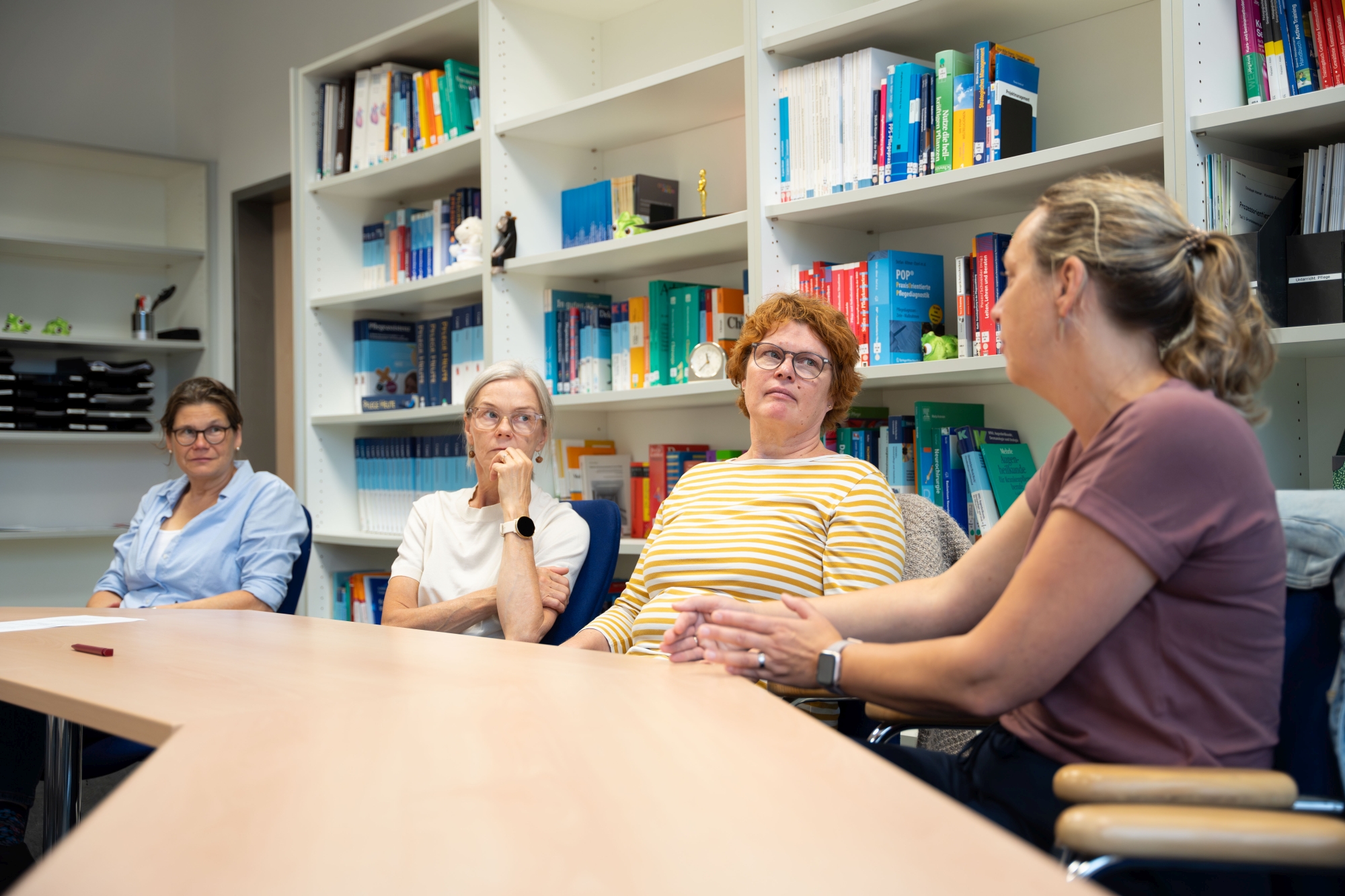Vier Frauen sitzen an einem Tisch in einem Raum mit Bücherregalen und sprechen miteinander. Zwei hören aufmerksam zu, während eine Teilnehmerin spricht.