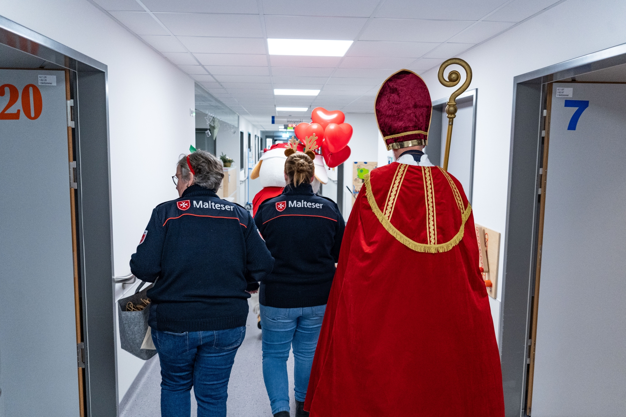 Zwei Malteser-Mitarbeiterinnen, das Maskottchen Eva des Verbunds mit roten Luftballons und der Nikolaus gehen gemeinsam einen Krankenhausflur entlang. Sie sind alle von hinten fotografiert.