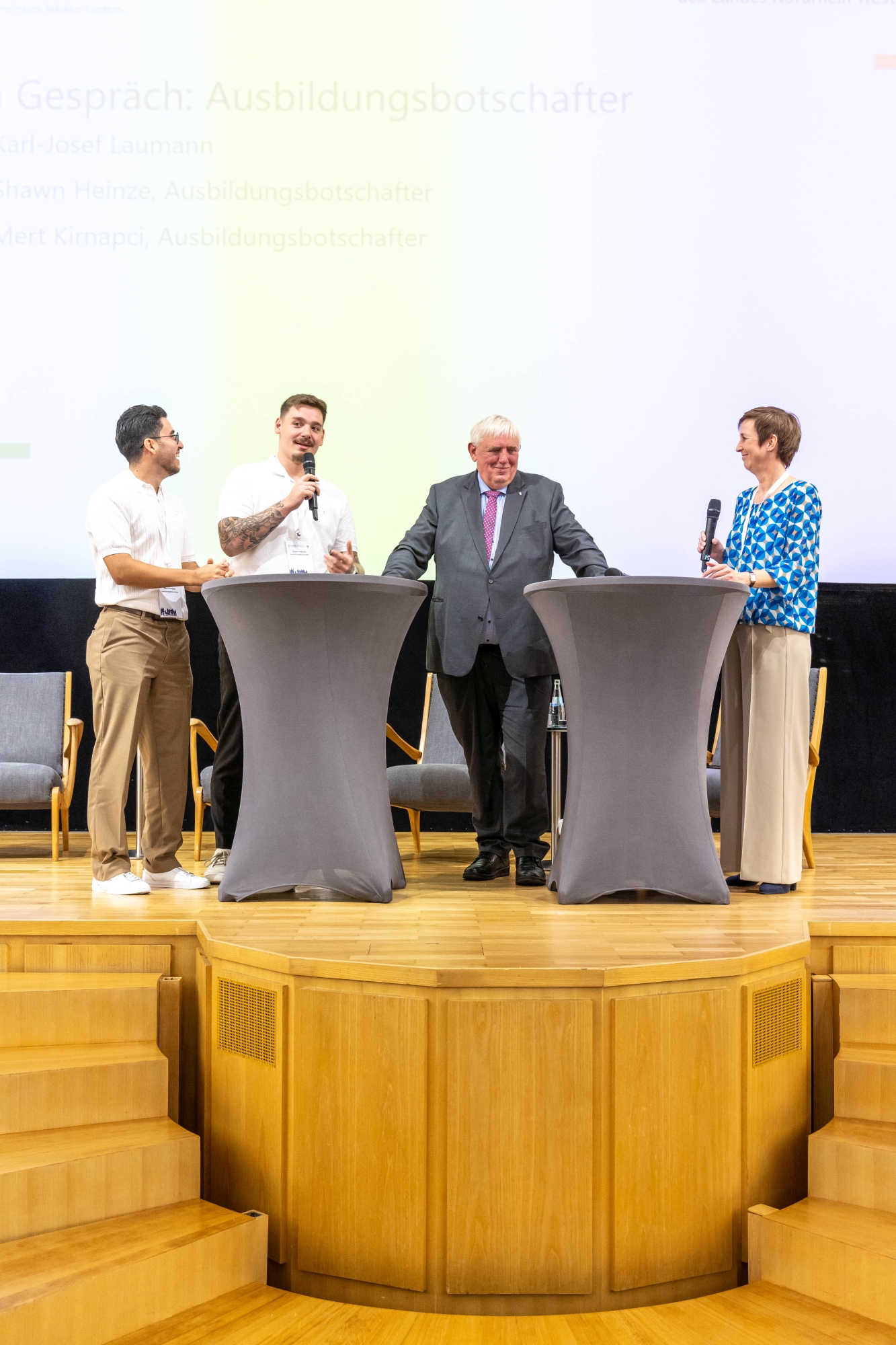 Podiumsgespräch beim Fachtag Zukunftsbündnis Pflege: Zwei junge Pflegeauszubildende im Gespräch mit NRW-Gesundheitsminister Karl-Josef Laumann und einer Moderatorin auf einer hellen Bühne mit zwei Stehtischen.
