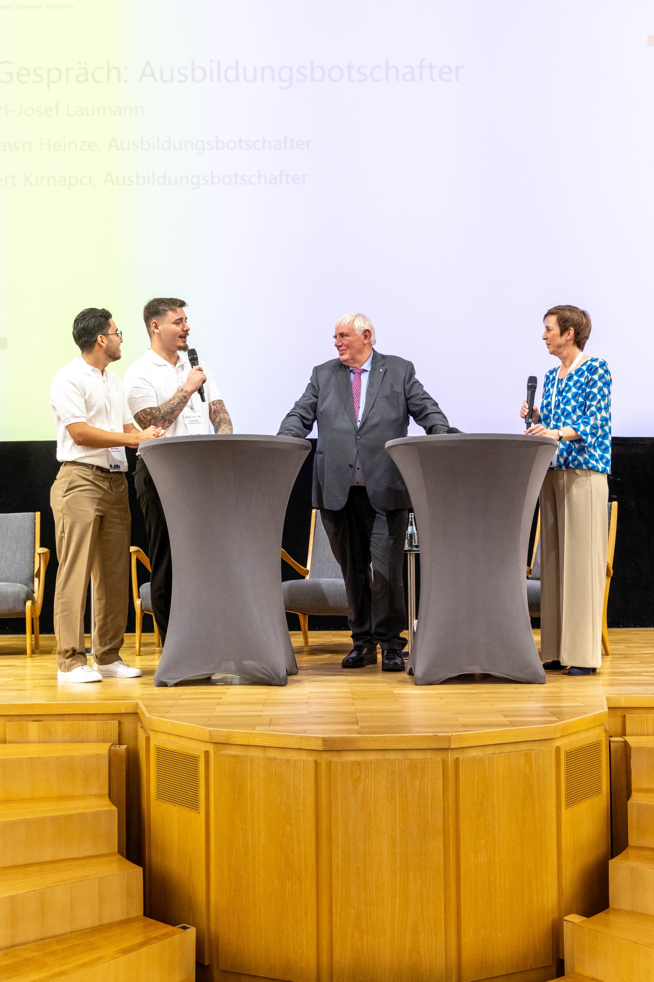 Auf dem Podium des Fachtags Zukunftsbündnis Pflege diskutieren zwei Auszubildende mit NRW-Gesundheitsminister Karl-Josef Laumann. Eine Moderatorin begleitet das Gespräch. Die Teilnehmenden stehen hinter zwei grauen Stehtischen auf einer Bühne.