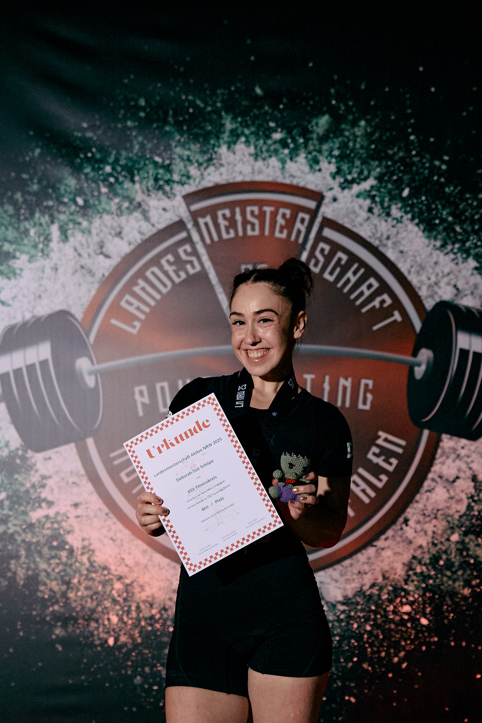 Die Kraftdreikämpferin Debby Schöpe steht lächelnd vor einer Wettkampfbühne und hält ihre Siegerurkunde sowie eine kleine Figur in den Händen. Im Hintergrund ist das Logo der Landesmeisterschaft im Powerlifting zu sehen.